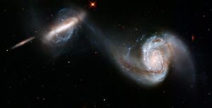A pair of merging galaxies. The galaxy on the right has a large, single spiral arm curving out from the core and around to above it, with very visible glowing dust and gas. The left galaxy is shaped like a long bar., surrounded by swirls of blue-white stars. A thin stream of whitish gas connects the two galaxies' cores. A few small stars and galaxies are scattered around the black background.